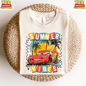 May include: Cream-colored t-shirt featuring a graphic of a red race car with the text "SUMMER VIBES". The design includes palm trees, colorful squares, and stars, evoking a summer theme. The shirt is made of a soft material.