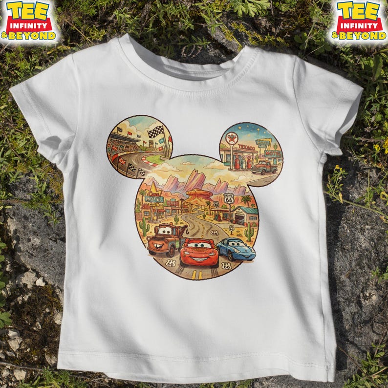 May include: White t-shirt featuring a colorful graphic of the Cars movie characters in a Mickey Mouse head shape. The design includes race tracks, desert landscapes, and Route 66 elements. The shirt has short sleeves and a crew neck.