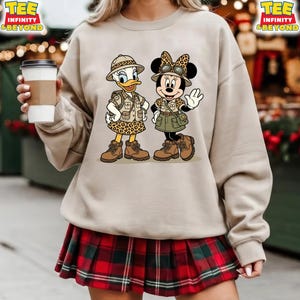 May include: Beige sweatshirt featuring a cartoon illustration of Daisy Duck and Minnie Mouse dressed in safari outfits. Daisy Duck wears a leopard print skirt and Minnie Mouse wears a green skirt. Both characters wear brown boots and hats. The sweatshirt has the text "TEE INFINITY & BEYOND".