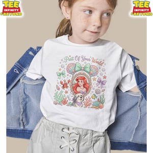 May include: White t-shirt featuring a colorful graphic of a mermaid with the text "Part Of Your World." The design includes seashells, a crab, and a dog. The shirt has a crew neck and short sleeves.