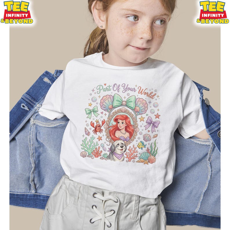 May include: White t-shirt featuring a colorful graphic of a mermaid with the text "Part Of Your World." The design includes seashells, a crab, and a dog. The shirt has a crew neck and short sleeves.