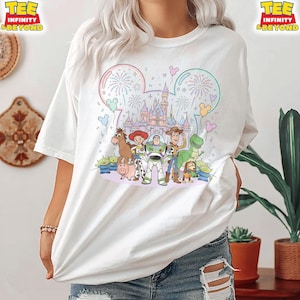 May include: White t-shirt featuring a colorful graphic of Disney's Toy Story characters in front of a castle. The design includes fireworks, Mickey Mouse ears, and the text "TEE INFINITY & BEYOND" on the top left and right.