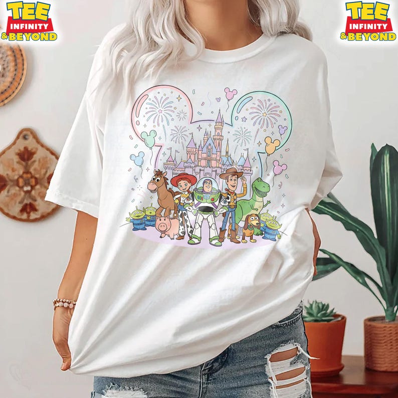 May include: White t-shirt featuring a colorful graphic of Disney's Toy Story characters in front of a castle. The design includes fireworks, Mickey Mouse ears, and the text "TEE INFINITY & BEYOND" on the top left and right.