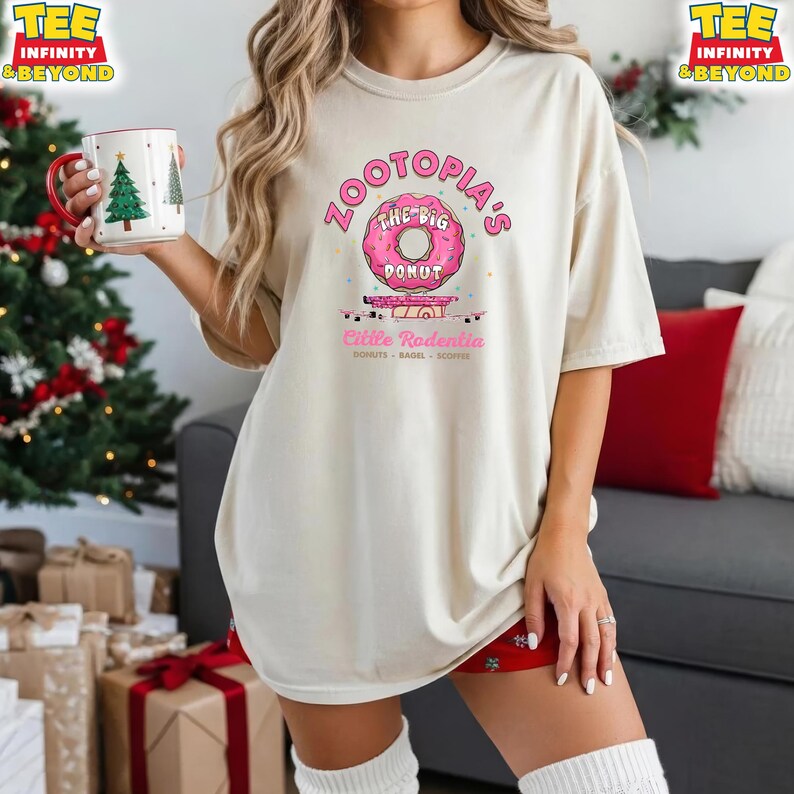 Puede incluir: Camiseta beige con el texto rosa "Zootopia's The Big Donut" y un gr&aacute;fico de donut rosa. Debajo, el texto "Little Rodentia". La persona lleva una taza con un dise&ntilde;o de &aacute;rbol de Navidad.