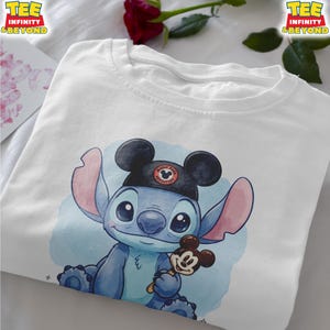 K&ouml;nnte beinhalten: Wei&szlig;es T-Shirt mit einer blauen Cartoon-Figur, die einen schwarzen Mickey-Mouse-Ohrenhut mit dem Text "Magic Park Adventure" tr&auml;gt. Die Figur h&auml;lt ein Mickey-Mouse-Eis am Stiel. Das T-Shirt hat den Text "TEE INFINITY & BEYOND".