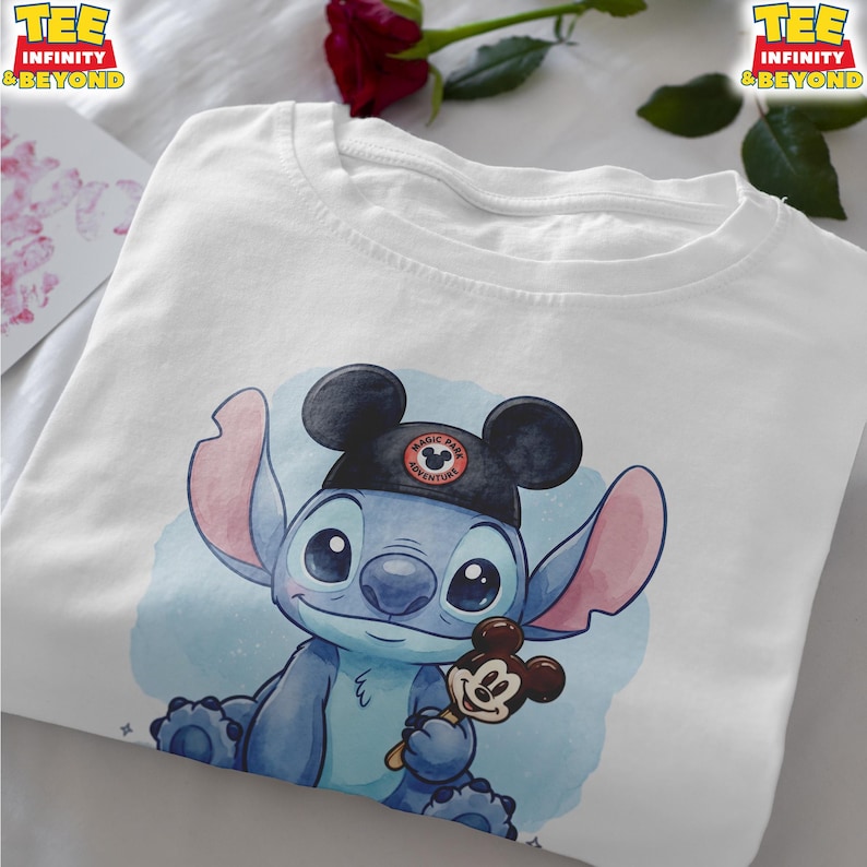 K&ouml;nnte beinhalten: Wei&szlig;es T-Shirt mit einer blauen Cartoon-Figur, die einen schwarzen Mickey-Mouse-Ohrenhut mit dem Text "Magic Park Adventure" tr&auml;gt. Die Figur h&auml;lt ein Mickey-Mouse-Eis am Stiel. Das T-Shirt hat den Text "TEE INFINITY & BEYOND".