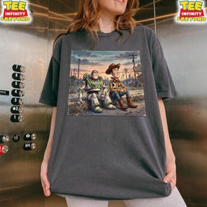 May include: Dark grey t-shirt with a colourful illustration of Buzz Lightyear and Woody from Toy Story. The image shows them sitting together in a city. The shirt has the text "TEE INFINITY & BEYOND".