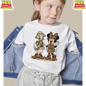 May include: White t-shirt featuring cartoon characters Donald Duck and Minnie Mouse in safari outfits. Donald Duck wears a khaki safari hat and vest, while Minnie Mouse sports a leopard print outfit. The shirt has the text "TEE INFINITY & BEYOND" in the upper corners.