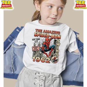 Op de afbeelding: Een wit t-shirt met een afbeelding van Spider-Man die door New York City slingert. De tekst luidt THE AMAZING SPIDER-MAN, SWING, NEW YORK CITY, SPRING INTO ACTION en 1962. Het ontwerp bevat een spinnenweb-achtergrond en een kleine cirkelvormige inzet.