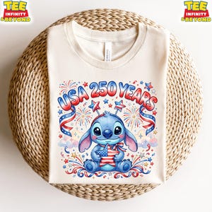 May include: Cream-colored t-shirt featuring a cartoon Stitch character holding a star-shaped American flag. The shirt has the text "USA 250 YEARS" in red, white, and blue, with fireworks and star accents. The shirt is displayed on a woven surface.