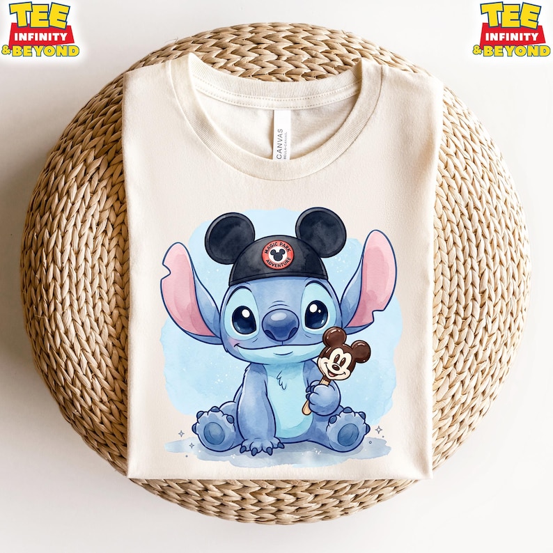 K&ouml;nnte beinhalten: Cremefarbenes T-Shirt mit einer blauen Cartoonfigur, die einen schwarzen Hut mit Mickey Mouse-Design tr&auml;gt. Die Figur h&auml;lt einen Mickey Mouse-Eis am Stiel. Das Shirt hat oben den Text "TEE INFINITY & BEYOND".