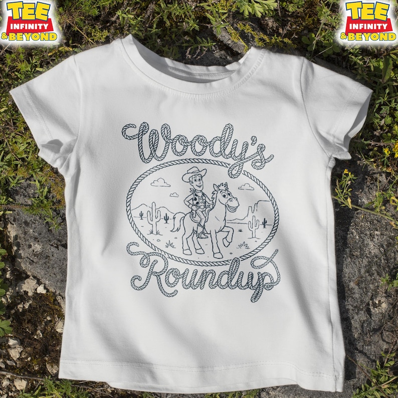 May include: A white short-sleeve t-shirt featuring a black line art graphic of a cowboy riding a horse within a rope-style oval frame. The text Woody's Roundup is written in a rope-style font above and below the central illustration.