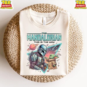 Może przedstawiać: Kremowy t-shirt z nadrukiem graficznym przedstawiającym postać Mandalorianina i małe zielone stworzenie. Tekst głosi THE MANDALORIAN THIS IS THE WAY. Wz&oacute;r zawiera planetę z pierścieniami, tr&oacute;jkątny kształt oraz gwiazdy w tle.