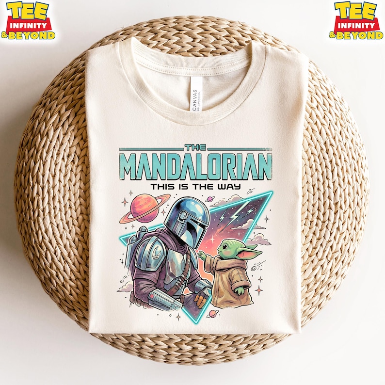 Może przedstawiać: Kremowy t-shirt z nadrukiem graficznym przedstawiającym postać Mandalorianina i małe zielone stworzenie. Tekst głosi THE MANDALORIAN THIS IS THE WAY. Wz&oacute;r zawiera planetę z pierścieniami, tr&oacute;jkątny kształt oraz gwiazdy w tle.