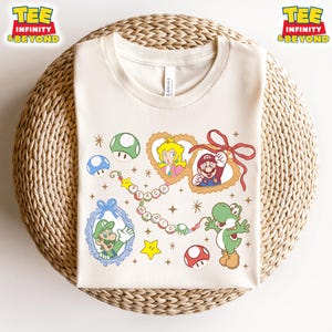 Puede incluir: Camiseta de color crema con personajes de dibujos animados de la serie Super Mario Bros. El diseño incluye retratos enmarcados de la Princesa Peach y Mario, junto con Yoshi y Luigi. Gráficos adicionales incluyen setas y estrellas.