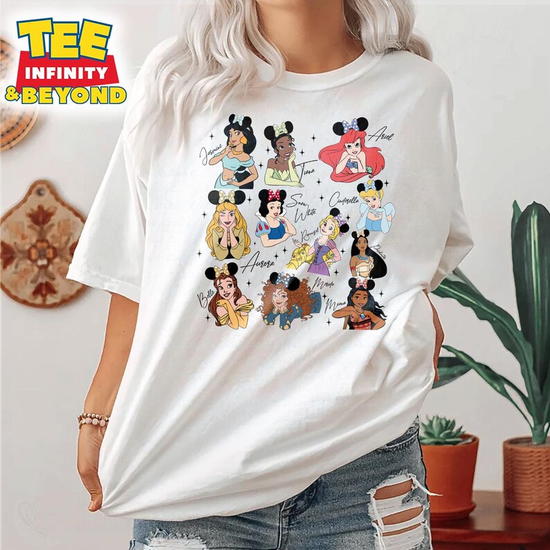 May include: White t-shirt with a graphic of Disney princesses wearing mouse ears. The princesses are: Belle, Aurora, Snow White, Cinderella, Tiana, Ariel, Jasmine, and Rapunzel. Each princess has her name written next to her.