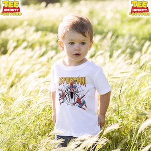 May include: A white t-shirt featuring a Spider-Man graphic. The design includes the text "SPIDER-MAN" in gold lettering, along with illustrations of Spider-Man and a spider web. The shirt is worn by a young child.