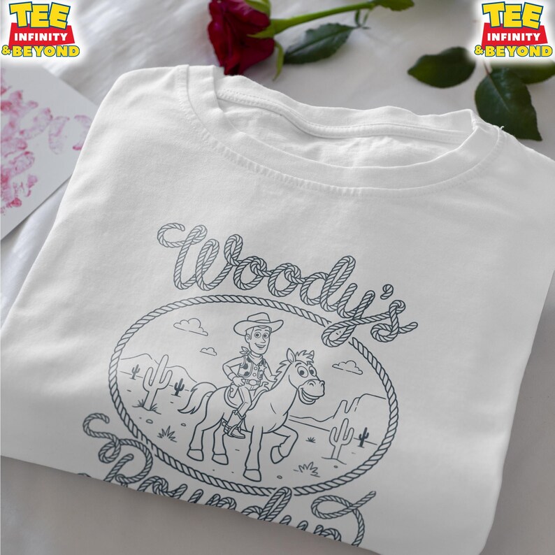 May include: A white t-shirt featuring a black line art graphic of a cowboy riding a horse inside a rope-style oval frame. The text "Woody's Roundup" is written in a rope-style font above and below the illustration, which includes desert cacti.