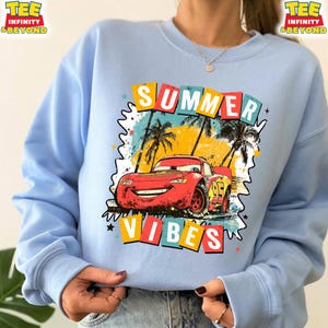 May include: Light blue sweatshirt featuring a graphic of a red race car with the number 95, palm trees, and the words "SUMMER VIBES" in colorful block letters. The sweatshirt has long sleeves and a crew neck.