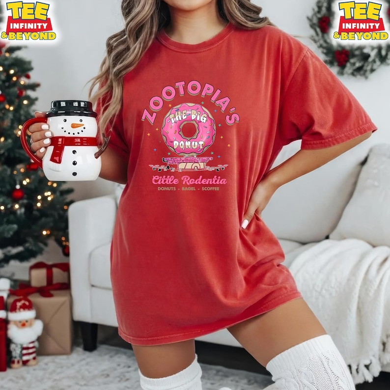 Puede incluir: Camiseta roja con el texto "Zootopia's The Big Donut" y un gr&aacute;fico de donut rosa. Debajo del donut est&aacute; el texto "Little Rodentia Donuts - Bagel - Scoffee". Se sostiene una taza de mu&ntilde;eco de nieve con una bufanda roja.