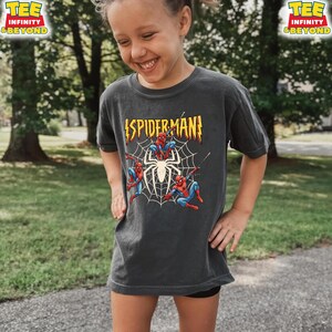 May include: Dark gray t-shirt featuring a Spider-Man graphic. The design includes the word "SPIDERMAN!" in yellow and red, along with illustrations of Spider-Man and a spiderweb. The shirt has short sleeves and a crew neck.
