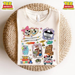 Puede incluir: Una camiseta blanca con un diseño colorido que presenta varios personajes y lugares de Disney. El diseño incluye a Mickey Mouse, Minnie Mouse, Woody, Buzz Lightyear y más. El texto "Hollywood Studios" y "Pizza Planet" también se incluye en el diseño.