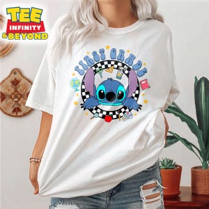 May include: White t-shirt featuring a cartoon character with the text "FIRST GRADE" in a circular design. The design includes school-themed elements like a globe, apple, and scissors. The shirt has short sleeves.