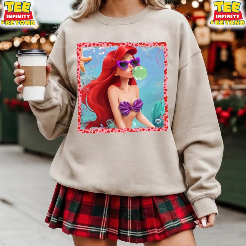 May include: Beige sweatshirt featuring a graphic of a mermaid with red hair, purple seashell top, and sunglasses blowing a bubble. The graphic is framed with a pink heart border. The words "TEE INFINITY & BEYOND" are printed on the top right and left.