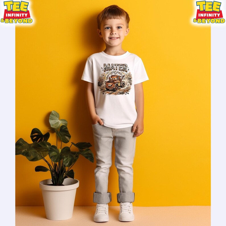 May include: A young child wearing a white t-shirt with a graphic of the character Mater from the movie Cars. The shirt has the word "MATER" above the image of the character. The child is wearing gray pants and white sneakers. The background is yellow.