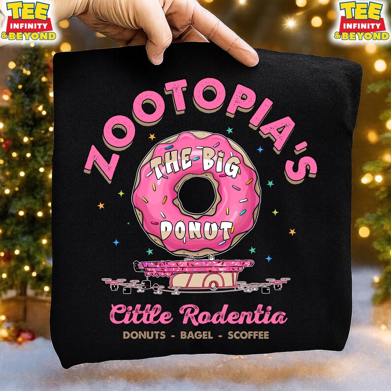 Puede incluir: Camiseta negra con un dise&ntilde;o de donut rosa. El texto dice "ZOOTOPIA'S THE BIG DONUT" y "Cittle Rodentia DONUTS - BAGEL - SCOFFEE". El dise&ntilde;o presenta un esquema de color rosa y blanco con estrellas.