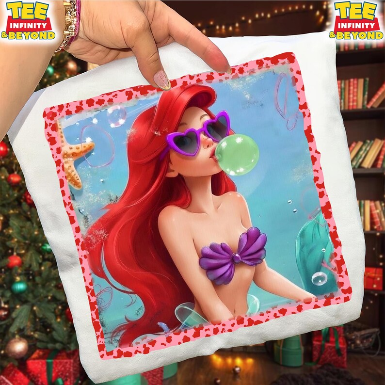 May include: White pillow with a colorful graphic of a mermaid wearing heart-shaped sunglasses and blowing a bubble. The mermaid has red hair and a purple seashell top. The pillow has a pink and red border and the text "TEE INFINITY & BEYOND".