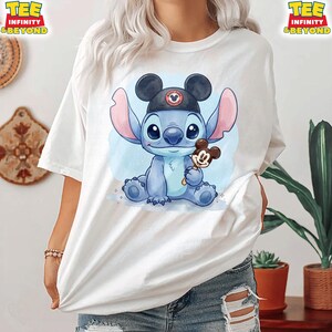K&ouml;nnte beinhalten: Wei&szlig;es T-Shirt mit einer blauen Cartoonfigur, die einen schwarzen Hut tr&auml;gt und ein Mickey Mouse-Eis am Stiel h&auml;lt. Die Figur sitzt auf einem hellblauen Hintergrund mit Aquarelleffekt. Das Shirt hat den Text "TEE INFINITY & BEYOND".