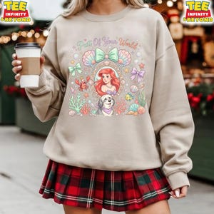 May include: Beige sweatshirt featuring a colorful graphic of Ariel from The Little Mermaid, surrounded by sea creatures and the text "Part Of Your World." The design includes a dog, shells, and bows.