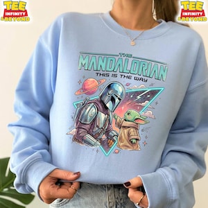 Może przedstawiać: Jasnoniebieska bluza z okrągłym dekoltem i nadrukiem graficznym przedstawiającym Mandalorianina oraz Grogu. Wz&oacute;r zawiera napis "THE MANDALORIAN THIS IS THE WAY" nad postaciami, na tle planety, gwiazd oraz neonowego tr&oacute;jkąta.