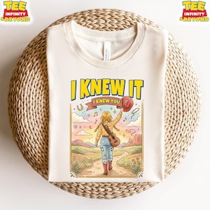 Puede incluir: Una camiseta color crema con una ilustraci&oacute;n gr&aacute;fica de una persona con cabello rubio en trenzas, vistiendo camisa a cuadros, jeans y botas rojas, caminando por un sendero con una guitarra y un sombrero. El texto dice I KNEW IT y I KNEW YOU.