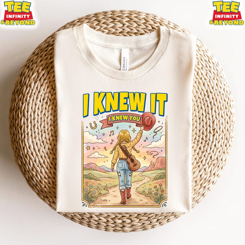 Puede incluir: Una camiseta color crema con una ilustraci&oacute;n gr&aacute;fica de una persona con cabello rubio en trenzas, vistiendo camisa a cuadros, jeans y botas rojas, caminando por un sendero con una guitarra y un sombrero. El texto dice I KNEW IT y I KNEW YOU.