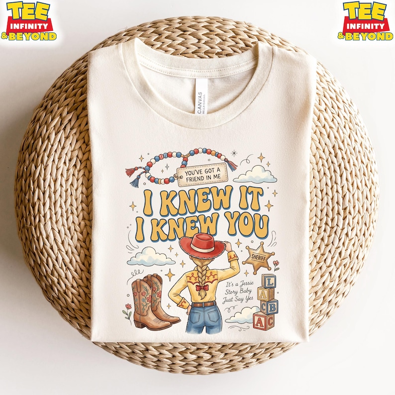 May include: A cream-colored t-shirt featuring a graphic print of a cowgirl with a braided ponytail, a red hat, and cowboy boots. The text reads: You've got a friend in me, I knew it, I knew you, It's a Jessie Story Baby Just Say Yes, and Sheriff.
