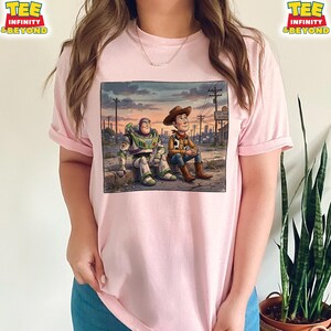 May include: Pale pink t-shirt with a graphic of Buzz Lightyear and Woody from Toy Story. The image shows them sitting together against a sunset backdrop with a cityscape. The shirt has the text "TEE INFINITY & BEYOND" on the top left and right.