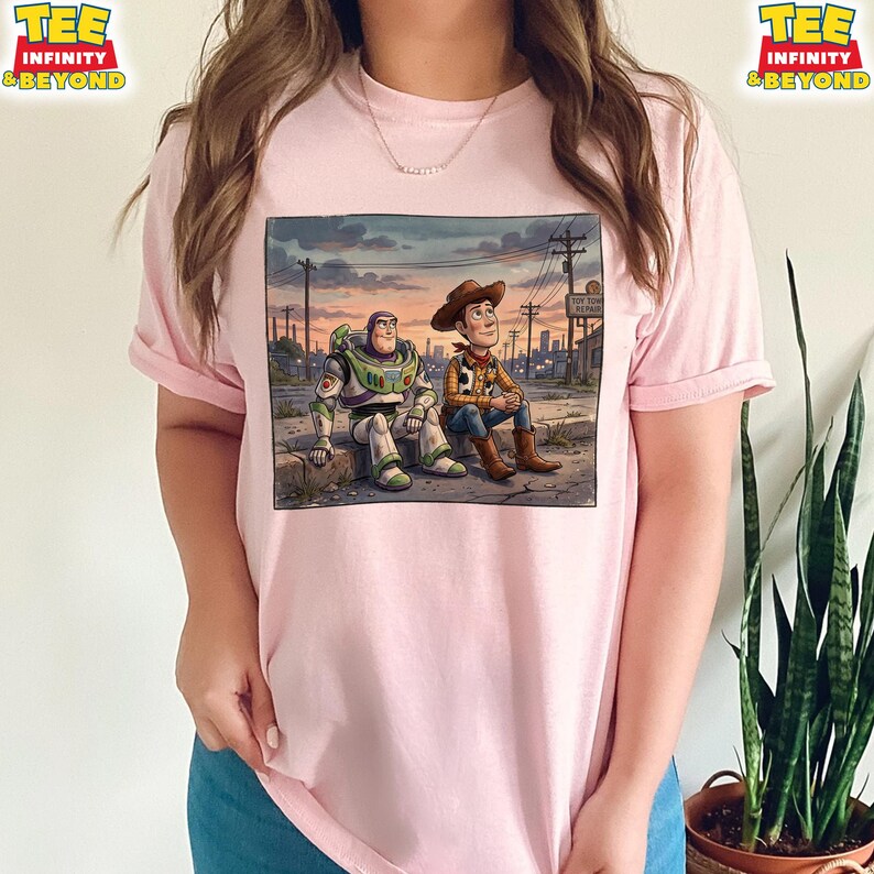 May include: Pale pink t-shirt with a graphic of Buzz Lightyear and Woody from Toy Story. The image shows them sitting together against a sunset backdrop with a cityscape. The shirt has the text "TEE INFINITY & BEYOND" on the top left and right.