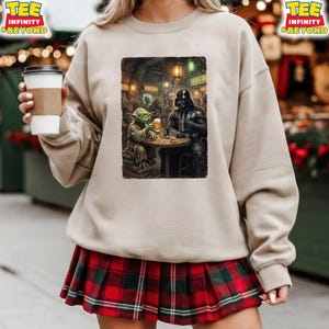 K&ouml;nnte beinhalten: Beiges Sweatshirt mit einem Aufdruck von Yoda und Darth Vader in einer Bar, jeder mit einem Bier. Das Bild spielt in einer schwach beleuchteten Bar mit h&auml;ngenden Laternen. Das Sweatshirt wird mit einem rot-gr&uuml;nen karierten Rock kombiniert.