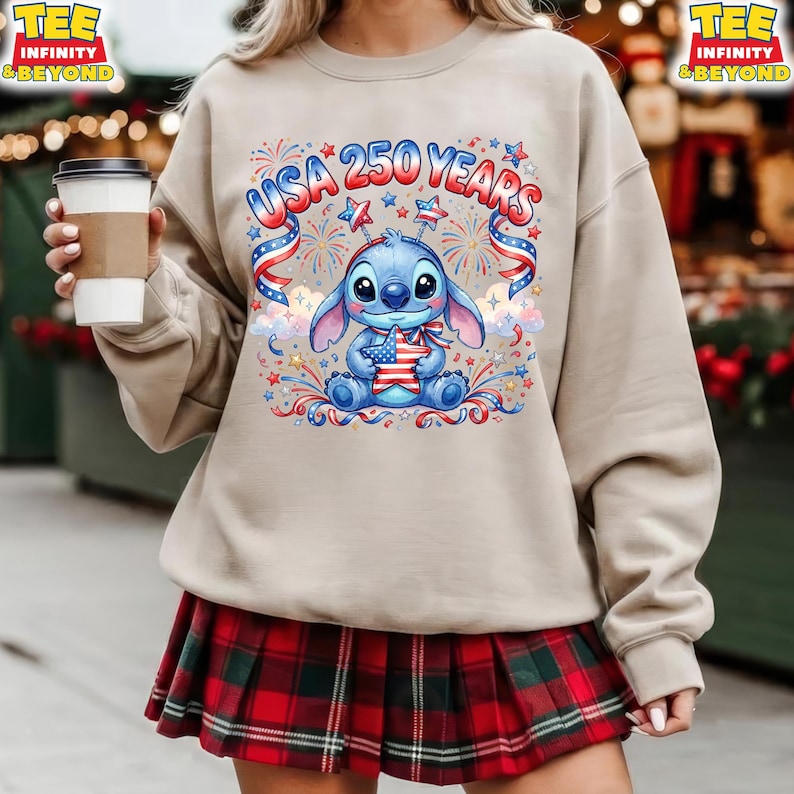 May include: Beige sweatshirt with a graphic of a blue cartoon character holding a star-shaped American flag, with the text "USA 250 YEARS" above. The design includes red, white, and blue fireworks and ribbons. The person is wearing a red and green plaid skirt.