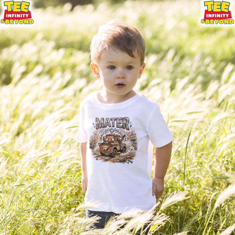 May include: A white t-shirt featuring a cartoon image of Mater from the Cars movie, with the name "MATER" above the image. The shirt is worn by a young child standing in a field of tall grass.