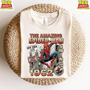 Op de afbeelding: Een cr&egrave;mekleurig t-shirt met ronde hals en een grafische print van Spider-Man die door New York City slingert. Het ontwerp bevat de tekst The Amazing Spider-Man, New York City, Spring Into Action en het jaartal 1962 in vintage stripstijl.