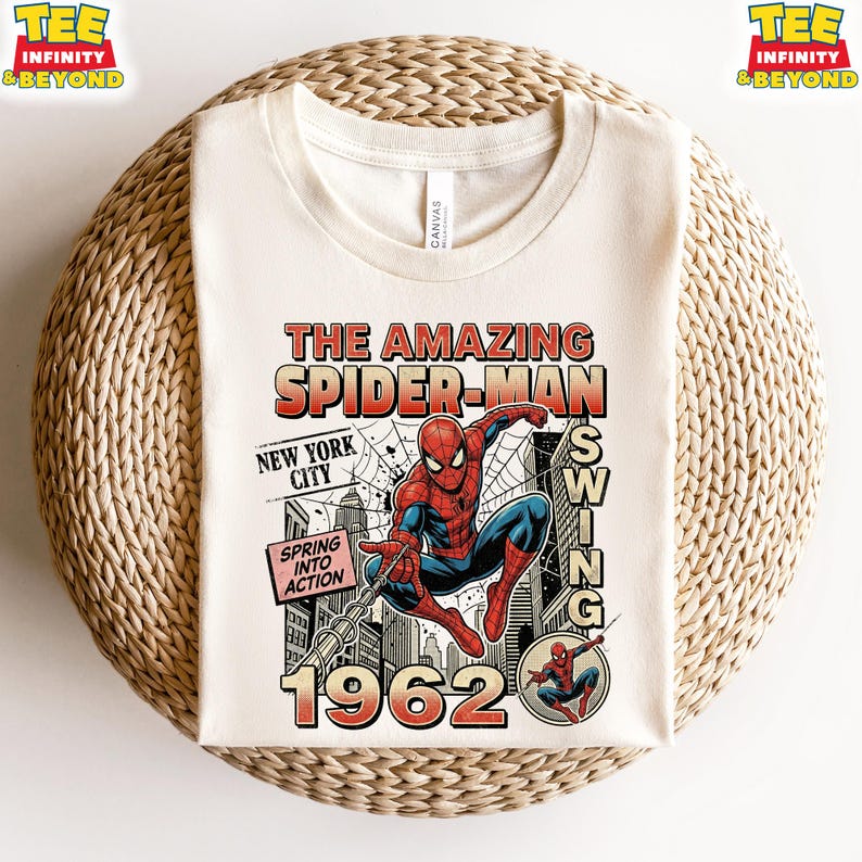 Op de afbeelding: Een cr&egrave;mekleurig t-shirt met ronde hals en een grafische print van Spider-Man die door New York City slingert. Het ontwerp bevat de tekst The Amazing Spider-Man, New York City, Spring Into Action en het jaartal 1962 in vintage stripstijl.