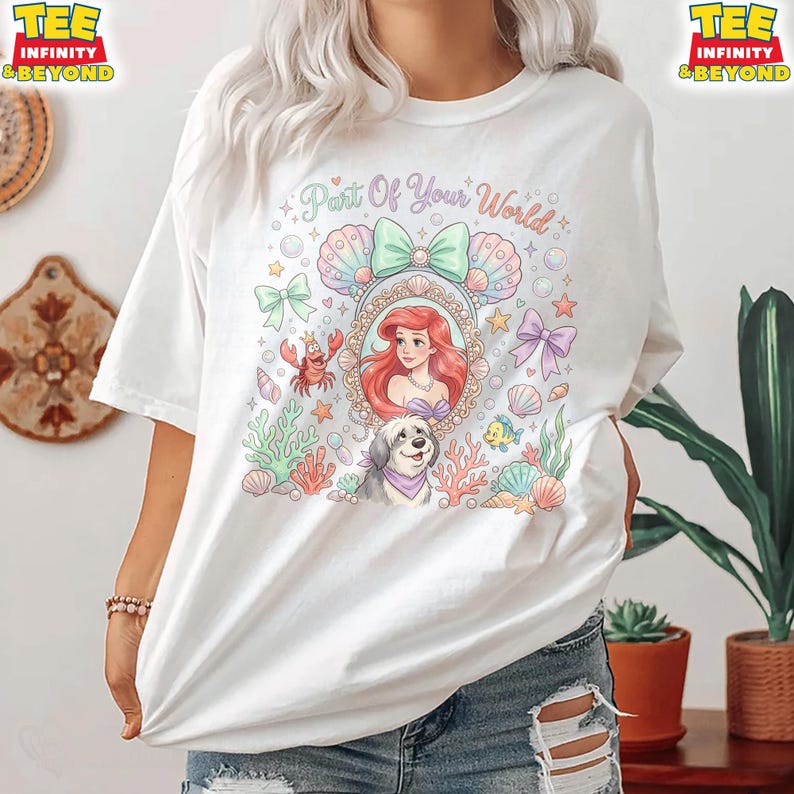 May include: White t-shirt featuring a colorful graphic of Ariel from The Little Mermaid, surrounded by seashells, bows, and a dog. The text "Part Of Your World" is arched above the image. The shirt has short sleeves and a relaxed fit.