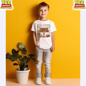 May include: A white short-sleeved t-shirt featuring a brown graphic print with a cowboy hat, boots, a cactus, and the text "I KNEW IT, I KNEW YOU" above the number 5, paired with light gray denim jeans and white sneakers.