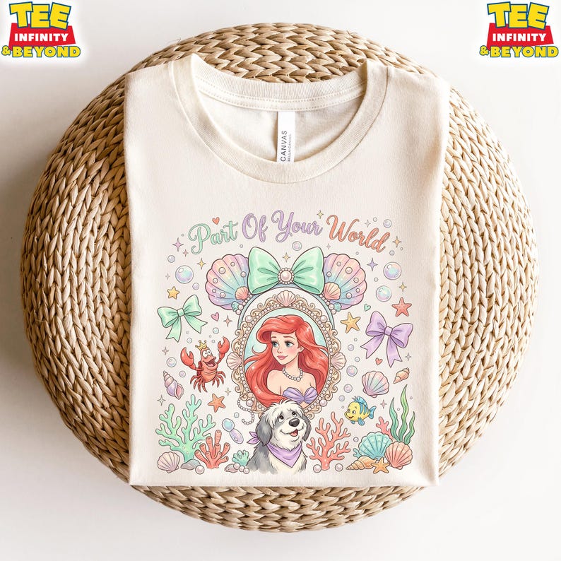May include: Cream-colored t-shirt featuring a colorful graphic of Ariel from The Little Mermaid, surrounded by sea creatures and the text "Part Of Your World." The design includes bows, shells, and a dog.