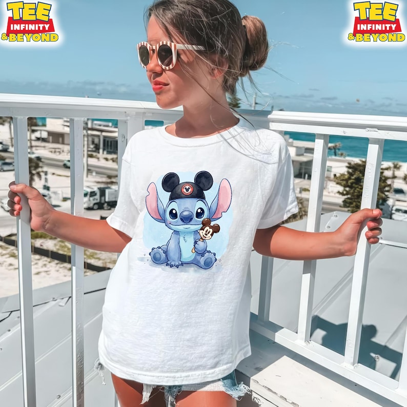 K&ouml;nnte beinhalten: Wei&szlig;es T-Shirt mit einer Cartoon-Illustration von Stitch, der einen Mickey Mouse-Hut tr&auml;gt und ein Mickey Mouse-Eis h&auml;lt. Das T-Shirt hat den Text "TEE INFINITY & BEYOND" in der oberen linken Ecke.