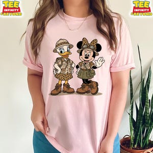 May include: Pink t-shirt featuring a cartoon illustration of Daisy Duck and Minnie Mouse dressed in safari outfits. Daisy Duck is wearing a safari hat and leopard print skirt. Minnie Mouse is wearing a safari hat with a bow and a green skirt.