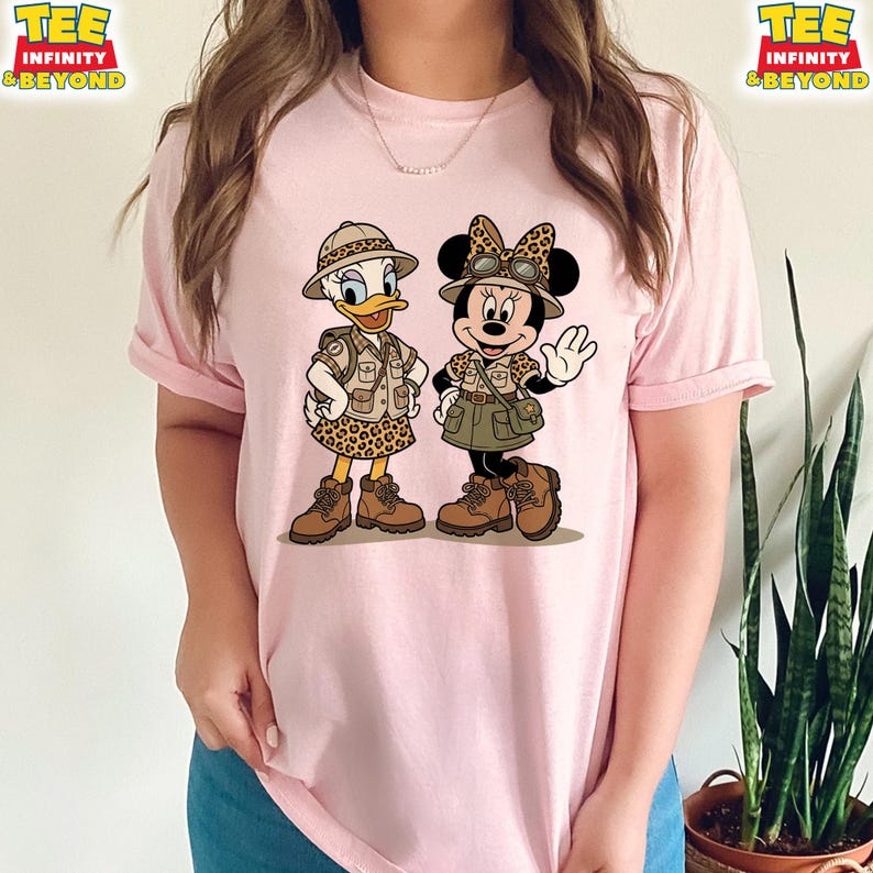 May include: Pink t-shirt featuring a cartoon illustration of Daisy Duck and Minnie Mouse dressed in safari outfits. Daisy Duck is wearing a safari hat and leopard print skirt. Minnie Mouse is wearing a safari hat with a bow and a green skirt.